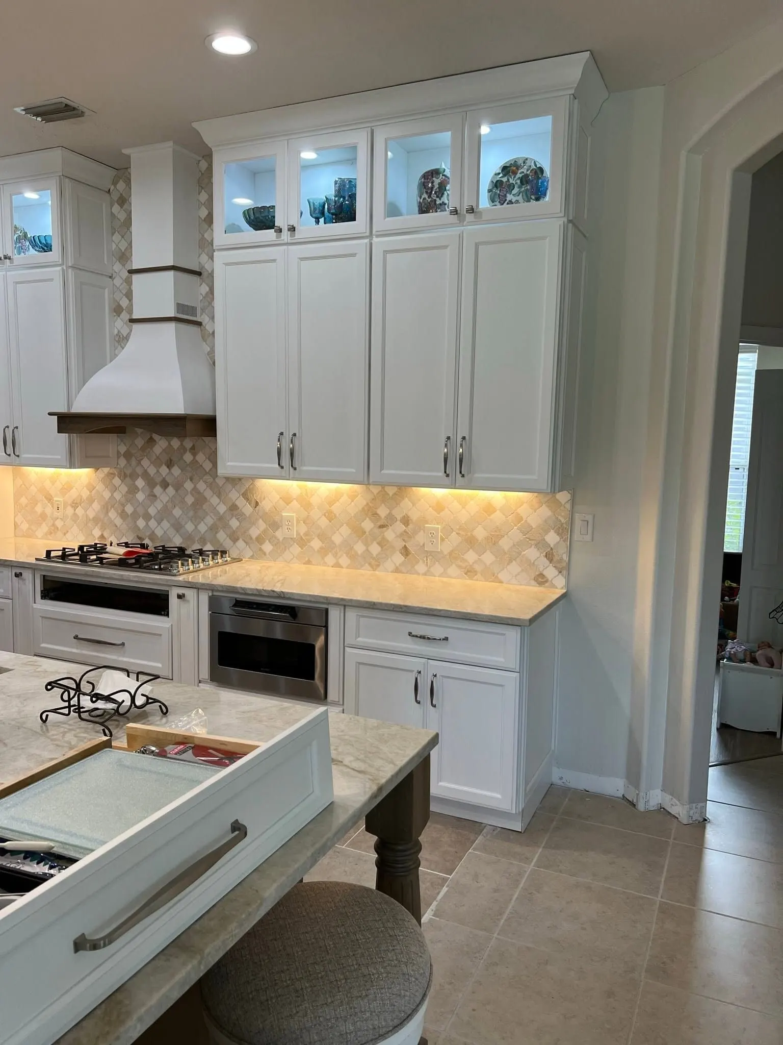 White kitchen with lit glass-front upper cabinets, a white range hood and a tile backsplash