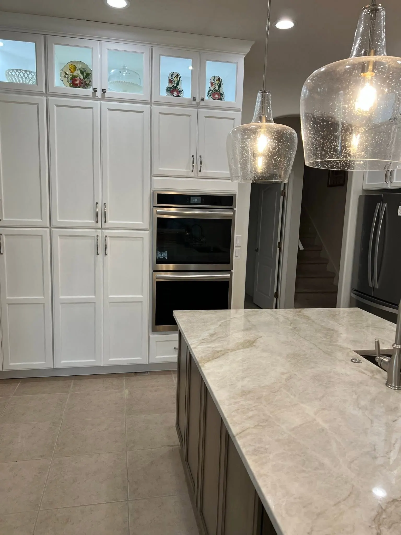 Floor-to-ceiling white pantry cabinets with glass-front tops and double wall ovens beside a stone-topped island